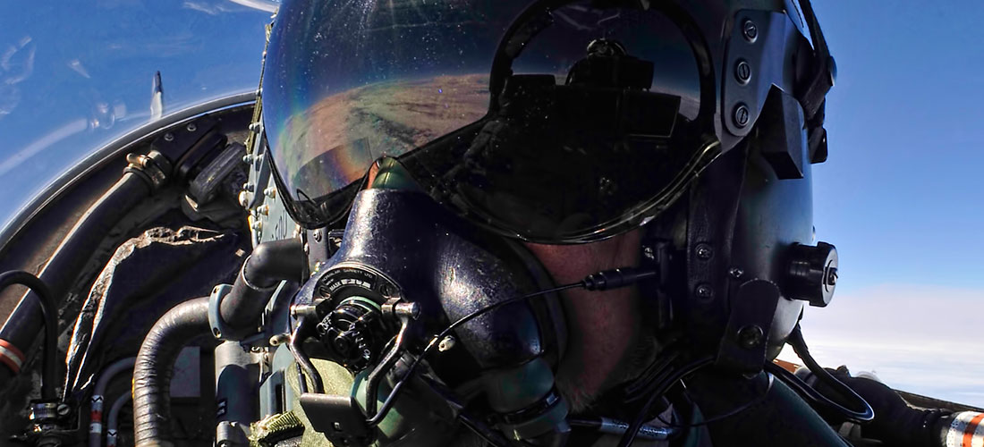 Test and Evaluation pilot in jet wearing helmet facing camera, above clouds with blue sky behind